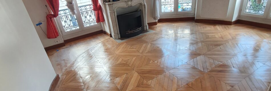 Pièce avec parquet clair au motif complexe, fenêtres avec rideaux rouges et voilages blancs, cheminée décorative et mur miroir. Murs beige/crème et plafond courbé. Porte en bois visible à gauche.