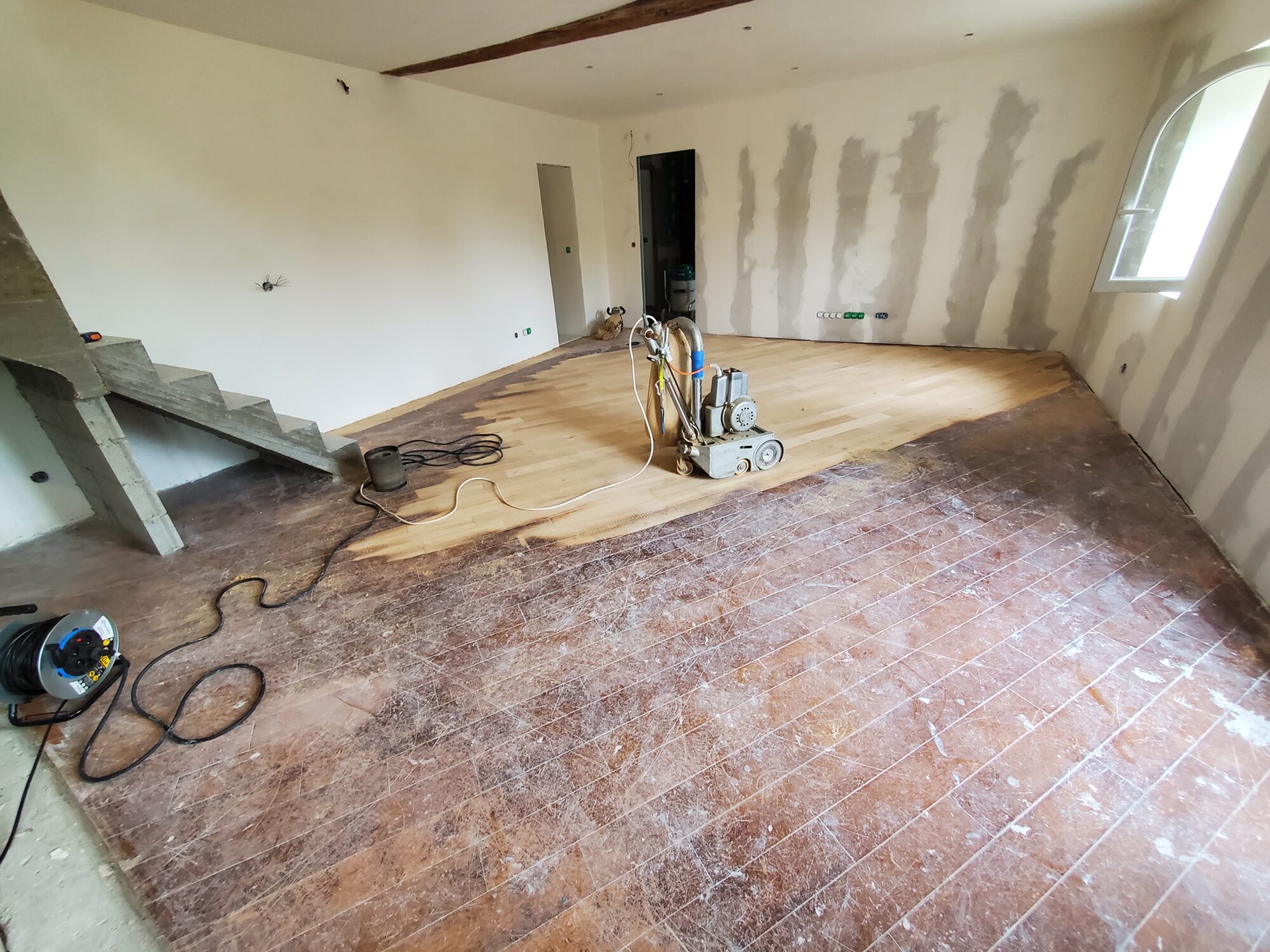 Pièce en rénovation avec sol en bois poncé, murs beige/crème et zones de plâtre inachevées. Escaliers en béton à l'arrière, câble d'extension visible, fenêtre en haut à droite.