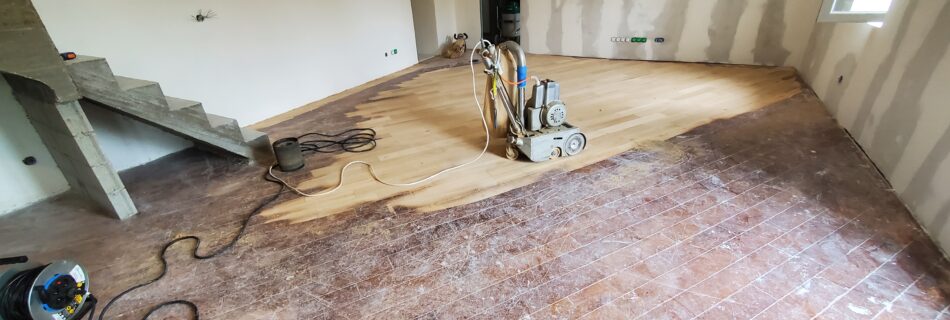 Pièce en rénovation avec sol en bois poncé, murs beige/crème et zones de plâtre inachevées. Escaliers en béton à l'arrière, câble d'extension visible, fenêtre en haut à droite.
