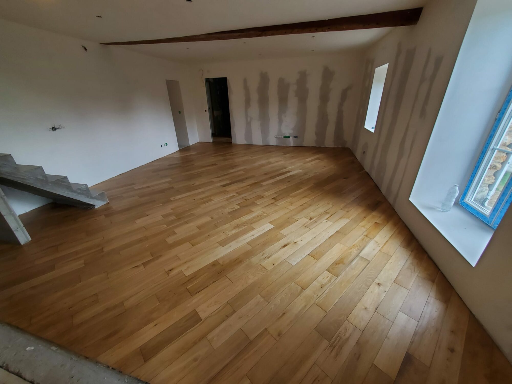 Pièce avec parquet en bois clair posé en diagonale, murs blancs en rénovation, escalier en béton à gauche, fenêtre bleue à droite, poutre en bois au plafond et porte non peinte à l'arrière.