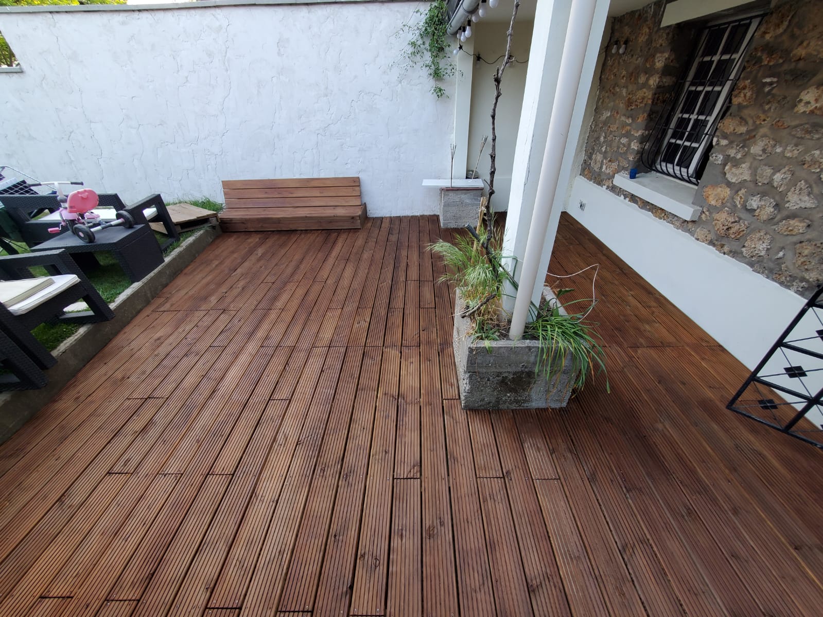 Terrasse en bois brun foncé avec plantes en pots, porche avec piliers blancs, mur en pierre à droite avec fenêtre sécurisée. Meubles de jardin sombres à gauche.