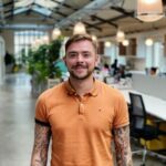 Un homme à la peau claire, cheveux bruns et barbe, sourit en polo orange avec des tatouages sur le bras. L'arrière-plan montre un espace de travail lumineux avec des plantes et des poutres métalliques.