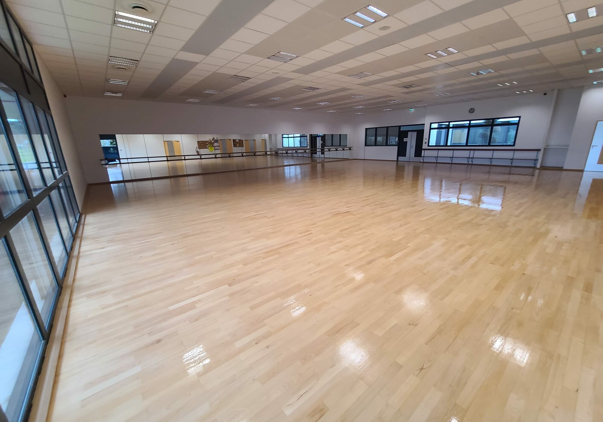 Une grande salle de danse lumineuse avec un sol en bois clair, des barres de ballet le long d'un mur et de grands miroirs. La pièce est vide et baignée de lumière naturelle.